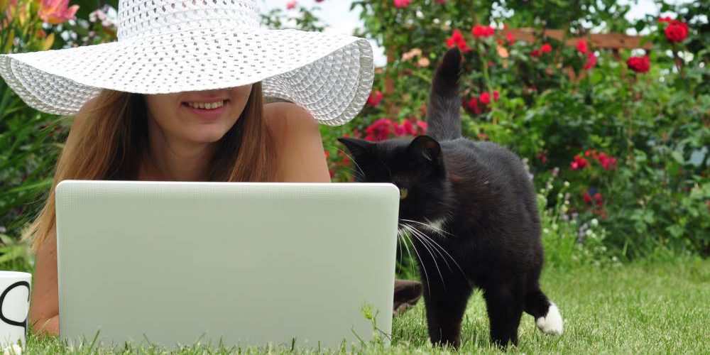 Ung kvinna i vit hatt med stort brätte ligger på gräset med sin bärbara dator. En svart katt står bredvid och kikar på skärmen. Kvinnan ler.
