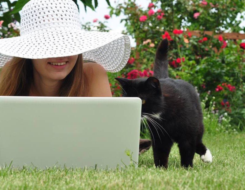 Ung kvinna i vit hatt med stort brätte ligger på gräset med sin bärbara dator. En svart katt står bredvid och kikar på skärmen. Kvinnan ler.
